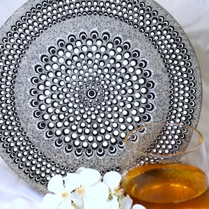 Puede incluir: Un plato decorativo gris con un patrón de puntos blancos y negros. El plato es redondo y tiene un diseño central de círculos concéntricos. Un vaso de líquido ámbar y flores blancas están en primer plano.