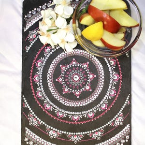 May include: A rectangular black slate board decorated with a hand-painted white, pink, and grey mandala dot pattern. A clear glass bowl filled with sliced red and yellow apples sits on the board, accompanied by a cluster of white flowers.