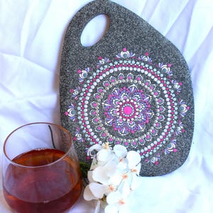 May include: A gray, hand-painted stone serving board with a mandala design in pink, purple, and white. A glass of amber liquid and white flowers are next to the board on a white surface.