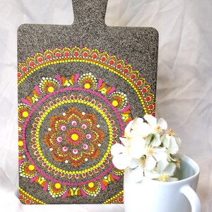 Puede incluir: Tabla de cortar gris con asa, con un vibrante diseño de mandala en puntos amarillos, rosas, naranjas y rojos. Una taza blanca con flores blancas está al lado.
