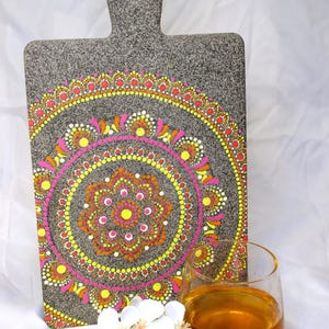 May include: A grey chopping board with a handle, decorated with a vibrant mandala pattern in pink, orange, and yellow. A glass of amber liquid and white flowers are in the foreground.