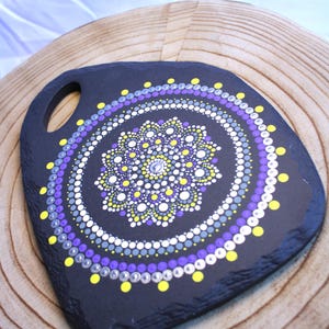 May include: A black slate board with a handle, featuring a hand-painted mandala design composed of concentric circles of dots in shades of purple, yellow, white, and grey. The centre of the mandala is accented with a small, clear, reflective gem.