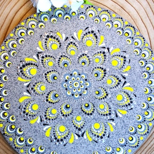 Puede incluir: Una piedra decorativa redonda pintada a mano con un diseño de mandala. La piedra gris presenta un patrón de puntos y formas florales amarillas, negras y blancas. La piedra está colocada sobre una superficie de madera y adornada con flores blancas.