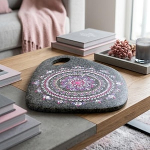 May include: A grey, oval decorative stone featuring a pink and purple mandala design. The stone has a handle and rests on a wooden coffee table. Other items on the table include books, a tray with flowers, and a candle.