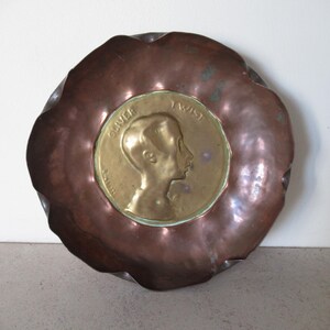 Puede incluir: Un plato de metal color cobre con un borde ondulado. El centro presenta un medallón dorado con el perfil de un joven y las palabras "OLIVER TWIST". El plato tiene un aspecto vintage.