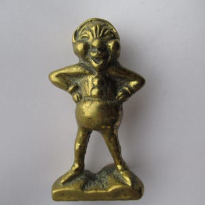 May include: A brass figurine of a smiling, cartoonish figure with arms akimbo. The figure has a round belly, small legs, and a hat-like headpiece. The figurine is standing on a small, irregular base.