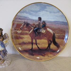Op de afbeelding: Een decoratief porseleinen bord met een gouden rand heeft een schilderij van twee mensen die op een paard door een woestijnlandschap rijden. Het bord staat op een wit oppervlak naast een porseleinen beeldje van een engel.