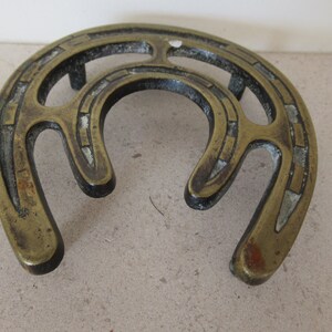 May include: A brass horseshoe-shaped pot holder with an intricate design. The holder has a raised center and two legs on either side.