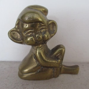 May include: A small, polished brass figurine depicting a seated character with large, rounded ears, a prominent nose, and closed eyes. The figure has a smooth, metallic gold-toned surface with visible signs of age and patina, showing a seated pose.