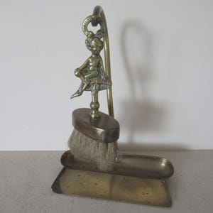 Può includere: Un set vintage di paletta e spazzola in ottone. La spazzola ha una testa rettangolare con setole di colore chiaro. Una piccola statuina di un cherubino seduto è appollaiata in cima a un manico curvo. La paletta è rettangolare con un bordo leggermente curvo.