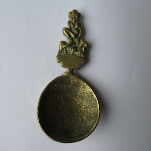 May include: A vintage brass spoon with a detailed mermaid figure atop the handle. The spoon's bowl is oval-shaped, and the handle features a seated mermaid with intricate detailing. The brass has a warm, aged patina, suggesting its antique nature.