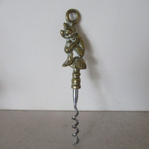 Puede incluir: Un sacacorchos de latón con una figura detallada de un gnomo sentado en un hongo. El sacacorchos es plateado y tiene un diseño en espiral.