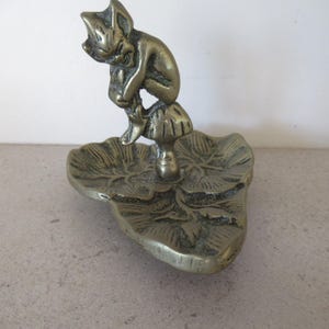 May include: A brass figurine of a whimsical creature resting on a mushroom, atop a leaf-shaped dish. The figurine has a detailed design and a vintage aesthetic. The dish is approximately 5 inches long.