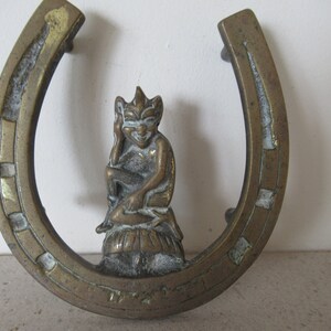 Könnte beinhalten: Ein Türstopper aus Messing in Form eines Hufeisens mit einer kleinen, detaillierten Gnomfigur, die obenauf sitzt. Das Hufeisen ist mit einem goldfarbenen Rand und weißen Akzenten verziert.