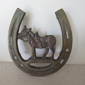 May include: Bronze-toned horseshoe-shaped decorative item with a donkey figure inside. The donkey has a saddle and bridle. The word "CLOVERLY" is inscribed at the bottom. The horseshoe has a rustic, aged appearance.