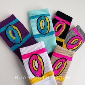 May include: A collection of colorful socks featuring a donut design. The socks come in various colors, including purple, teal, black, gray, white, and tan. Each sock has a donut graphic with a pink, yellow, and blue color scheme. The socks have striped accents.