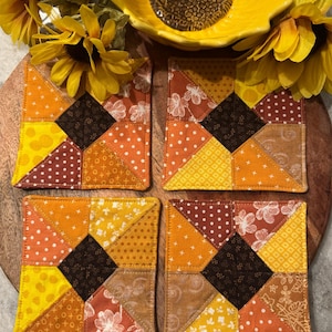 May include: Four square fabric coasters with a patchwork design in shades of yellow, orange, and brown. Each coaster features a central dark brown square surrounded by triangles of patterned fabric. The coasters are arranged on a wooden surface, with a yellow sunflower-shaped dish and artificial sunflowers.