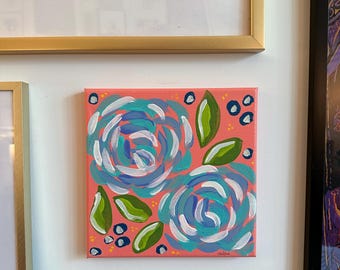 May include: Square canvas art featuring two abstract blue and white roses with green leaves and small blue dots on a coral background. The artwork is signed in the bottom right corner.