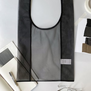 May include: A semi-transparent black tote bag with a rounded top and black trim. The bag is rectangular with a label attached. A notebook, pen, and glasses are next to the bag.