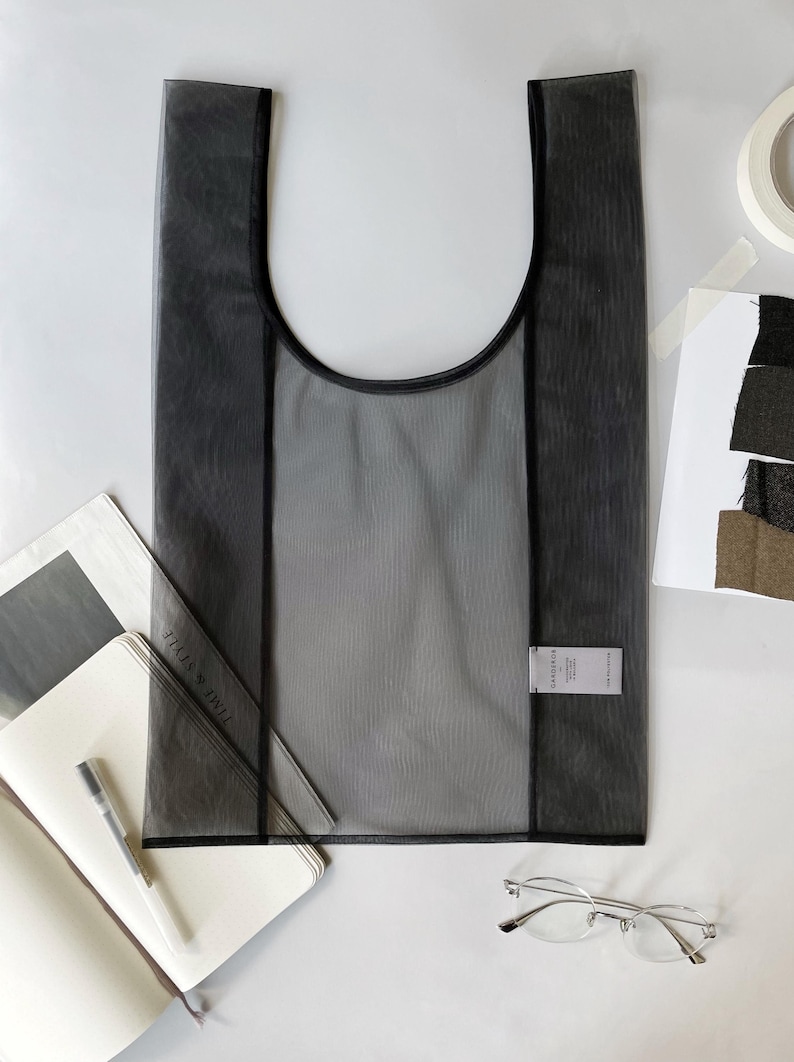 May include: A semi-transparent black tote bag with a rounded top and black trim. The bag is rectangular with a label attached. A notebook, pen, and glasses are next to the bag.