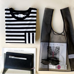 May include: A black and white striped t-shirt with a pocket, a black mesh tote bag with sunglasses and a magazine, and a black notebook with a pen. The magazine has Japanese text.