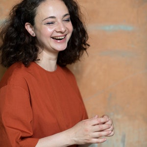 May include: A woman with curly brown hair is laughing while wearing a rust-coloured, short-sleeved shirt.