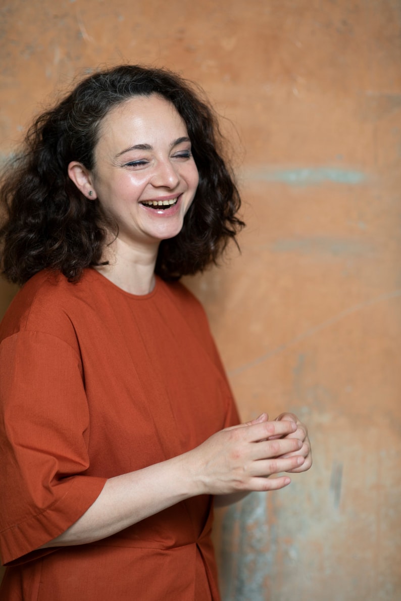 May include: A woman with curly brown hair is laughing while wearing a rust-coloured, short-sleeved shirt.