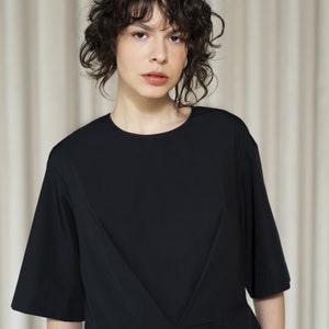 May include: A woman with curly dark hair is wearing a black short-sleeved dress with a V-shaped neckline. The dress has a pleated detail in the front.