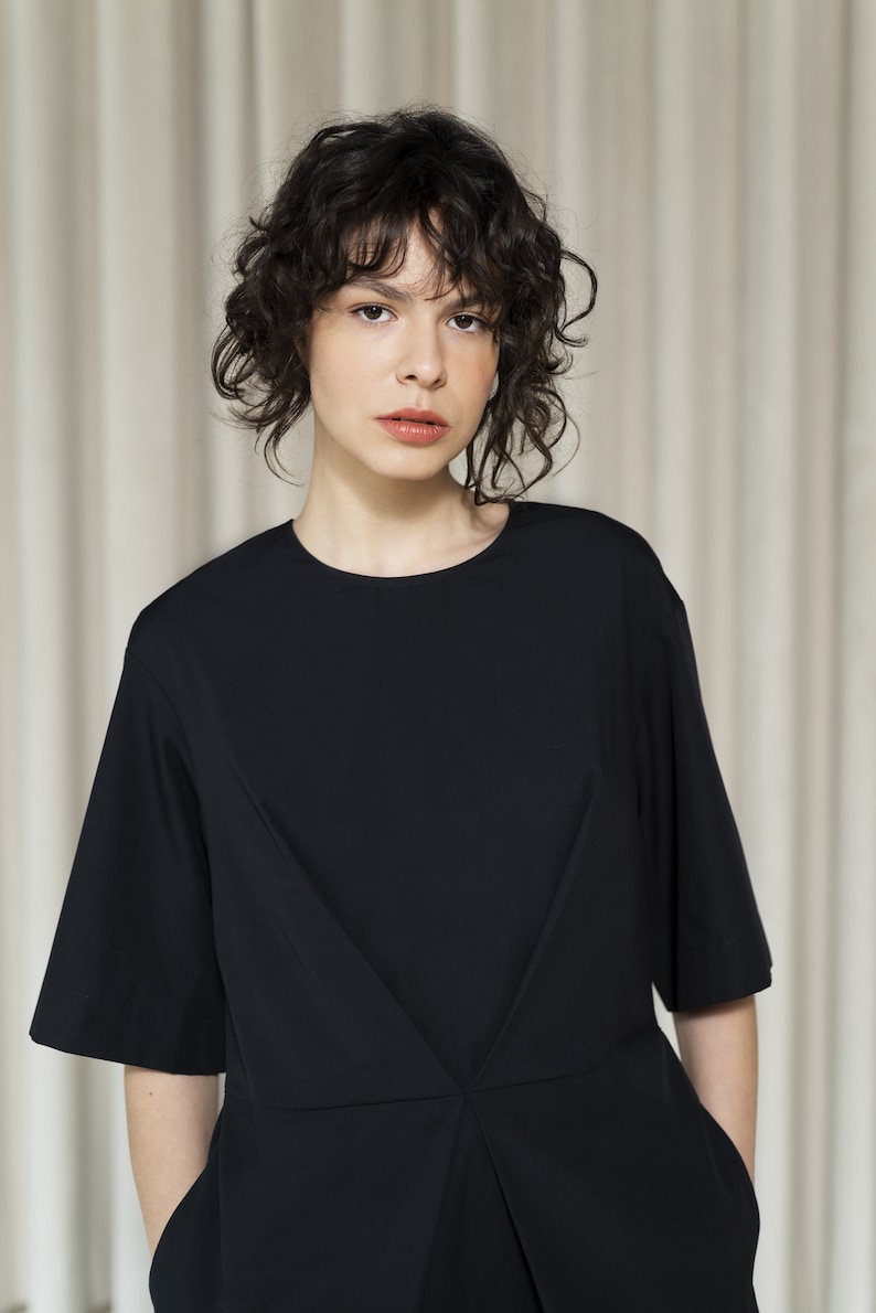 May include: A woman with curly dark hair is wearing a black short-sleeved dress with a V-shaped neckline. The dress has a pleated detail in the front.