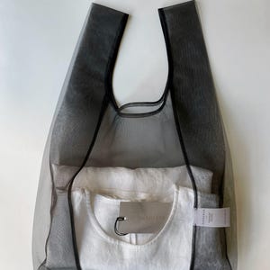 May include: A black mesh tote bag with a white t-shirt inside. The bag has two handles and a rectangular shape. A tag is attached to the t-shirt. The bag and t-shirt are on a white background.