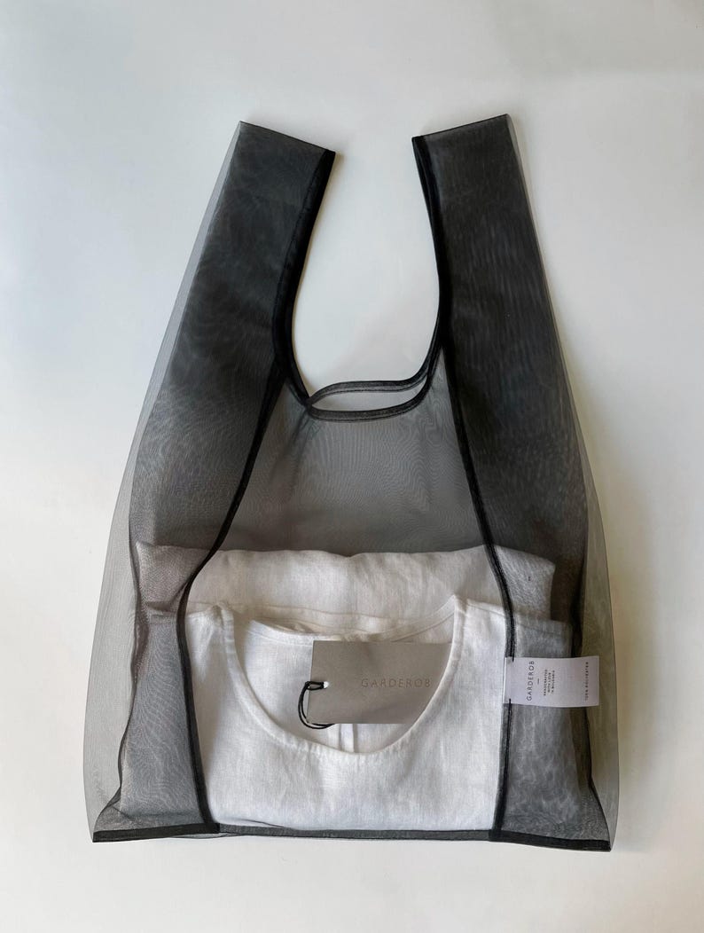 May include: A black mesh tote bag with a white t-shirt inside. The bag has two handles and a rectangular shape. A tag is attached to the t-shirt. The bag and t-shirt are on a white background.