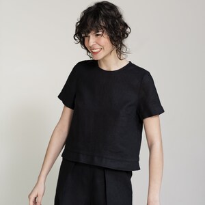 K&ouml;nnte beinhalten: Eine Frau tr&auml;gt ein schwarzes Leinenoberteil und eine passende Shorts. Das Oberteil hat kurze &Auml;rmel und einen runden Ausschnitt. Die Shorts haben eine gefaltete Vorderseite und sind knapp &uuml;ber dem Knie.