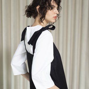 K&ouml;nnte beinhalten: Ein schwarzes Leinenkleid mit einem wei&szlig;en Hemd darunter. Das Kleid hat d&uuml;nne schwarze Tr&auml;ger, die an den Schultern zu Schleifen gebunden werden.
