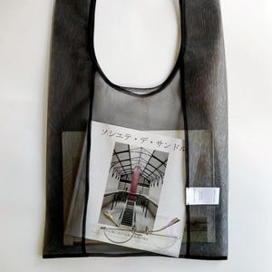 May include: A sheer black tote bag with a magazine, eyeglasses, and a small tag inside. The magazine has a photo of a building with a red structure. The eyeglasses have a thin, gold frame.