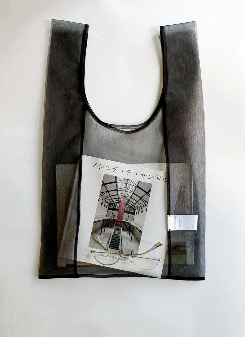 May include: A sheer black tote bag with a magazine, eyeglasses, and a small tag inside. The magazine has a photo of a building with a red structure. The eyeglasses have a thin, gold frame.