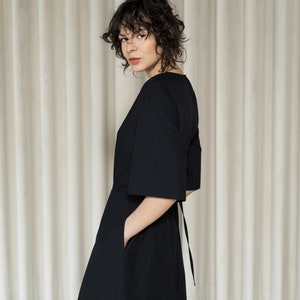 May include: A woman wearing a black, knee-length dress with a V-neck and a tie at the back. The dress has a loose fit and pockets on the sides.
