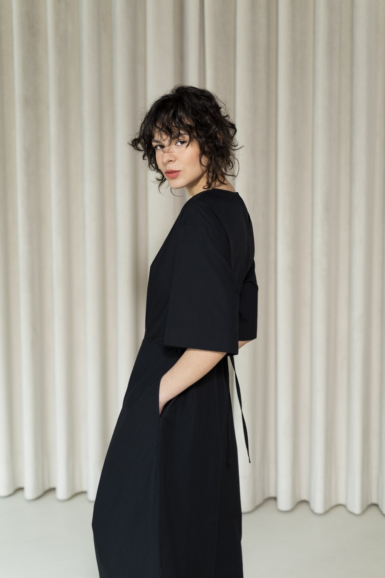 May include: A woman wearing a black, knee-length dress with a V-neck and a tie at the back. The dress has a loose fit and pockets on the sides.