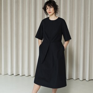 May include: A black midi dress with short sleeves and a cinched waist. The dress has pockets on both sides. The model is wearing black sandals.
