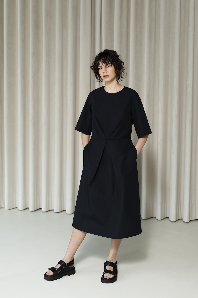 May include: A black midi dress with short sleeves and a cinched waist. The dress has pockets on both sides. The model is wearing black sandals.