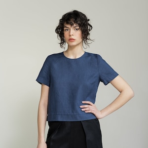 K&ouml;nnte beinhalten: Eine Frau tr&auml;gt ein blaues Leinenoberteil mit kurzen &Auml;rmeln und schwarze Leinenhosen. Sie steht mit der Hand auf der H&uuml;fte und blickt in die Kamera.