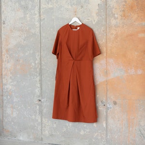 May include: A rust-coloured midi dress with short sleeves and a simple, slightly gathered design. The dress is hanging on a white hanger against a concrete wall.