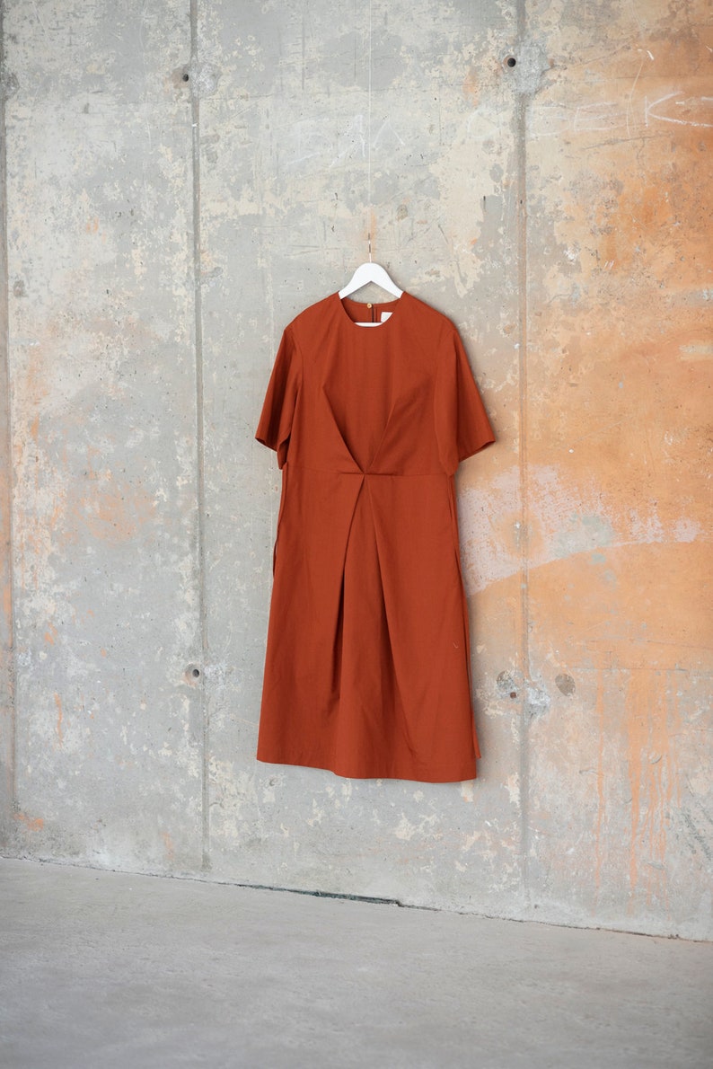 May include: A rust-coloured midi dress with short sleeves and a simple, slightly gathered design. The dress is hanging on a white hanger against a concrete wall.