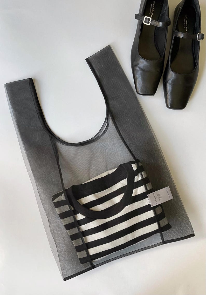 May include: A black mesh tote bag with a black and white striped shirt inside, and a pair of black leather Mary Jane style shoes with square toes. The items are arranged on a white surface.