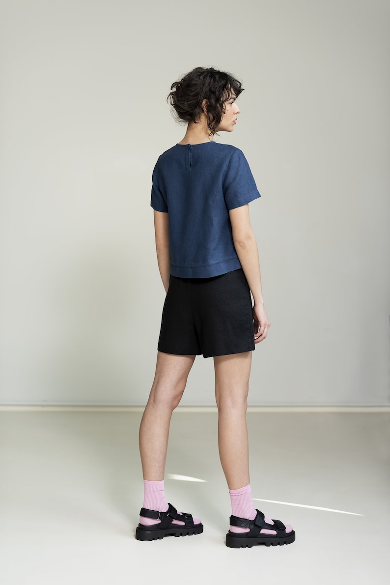 K&ouml;nnte beinhalten: Eine Frau tr&auml;gt ein dunkelblaues Leinenoberteil mit kurzen &Auml;rmeln und schwarze Leinenhosen. Sie tr&auml;gt rosa Socken und schwarze Sandalen.