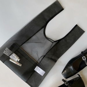 May include: A black mesh tote bag with a black interior pocket, containing a tube of cream and a lipstick. Two black leather Mary Jane style shoes are next to the bag. The items are on a white surface.