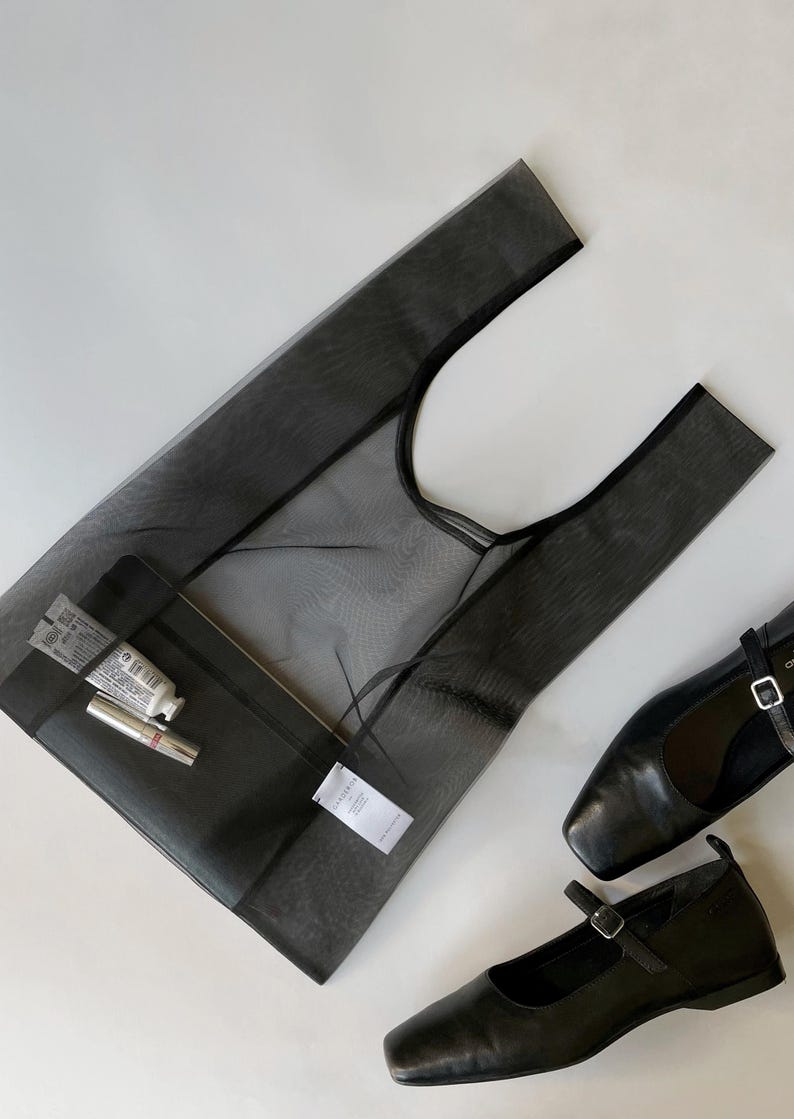 May include: A black mesh tote bag with a black interior pocket, containing a tube of cream and a lipstick. Two black leather Mary Jane style shoes are next to the bag. The items are on a white surface.
