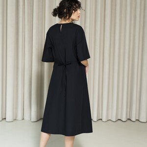 May include: A black midi dress with a tie at the waist. The dress has a round neckline and short sleeves. The model is wearing black sandals.