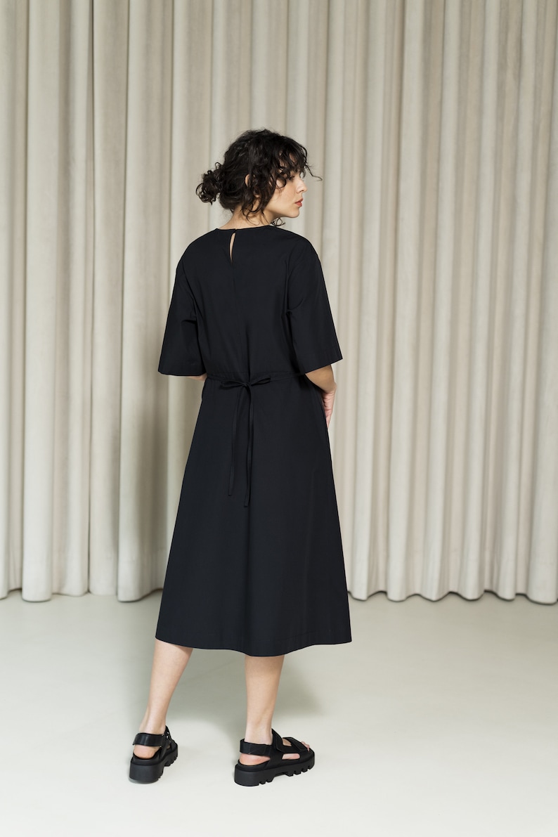 May include: A black midi dress with a tie at the waist. The dress has a round neckline and short sleeves. The model is wearing black sandals.
