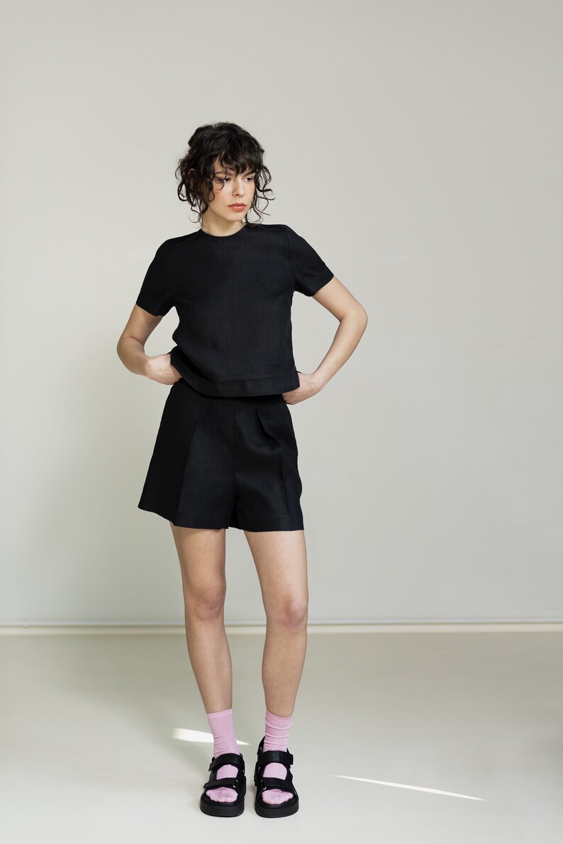 K&ouml;nnte beinhalten: Eine Frau tr&auml;gt ein schwarzes Leinenoberteil und eine passende Shorts. Sie tr&auml;gt rosa Socken und schwarze Sandalen.