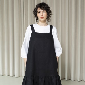 K&ouml;nnte beinhalten: Ein schwarzes Leinenkleid mit R&uuml;schen am Saum und d&uuml;nnen Tr&auml;gern. Das Kleid wird &uuml;ber einem wei&szlig;en T-Shirt getragen.